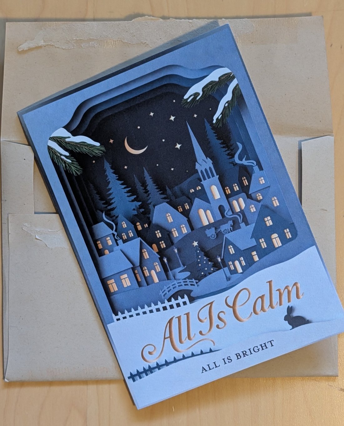 A layered papercut Christmas card, All Is Calm, resting on its kraft mailing envelope on a wooden table