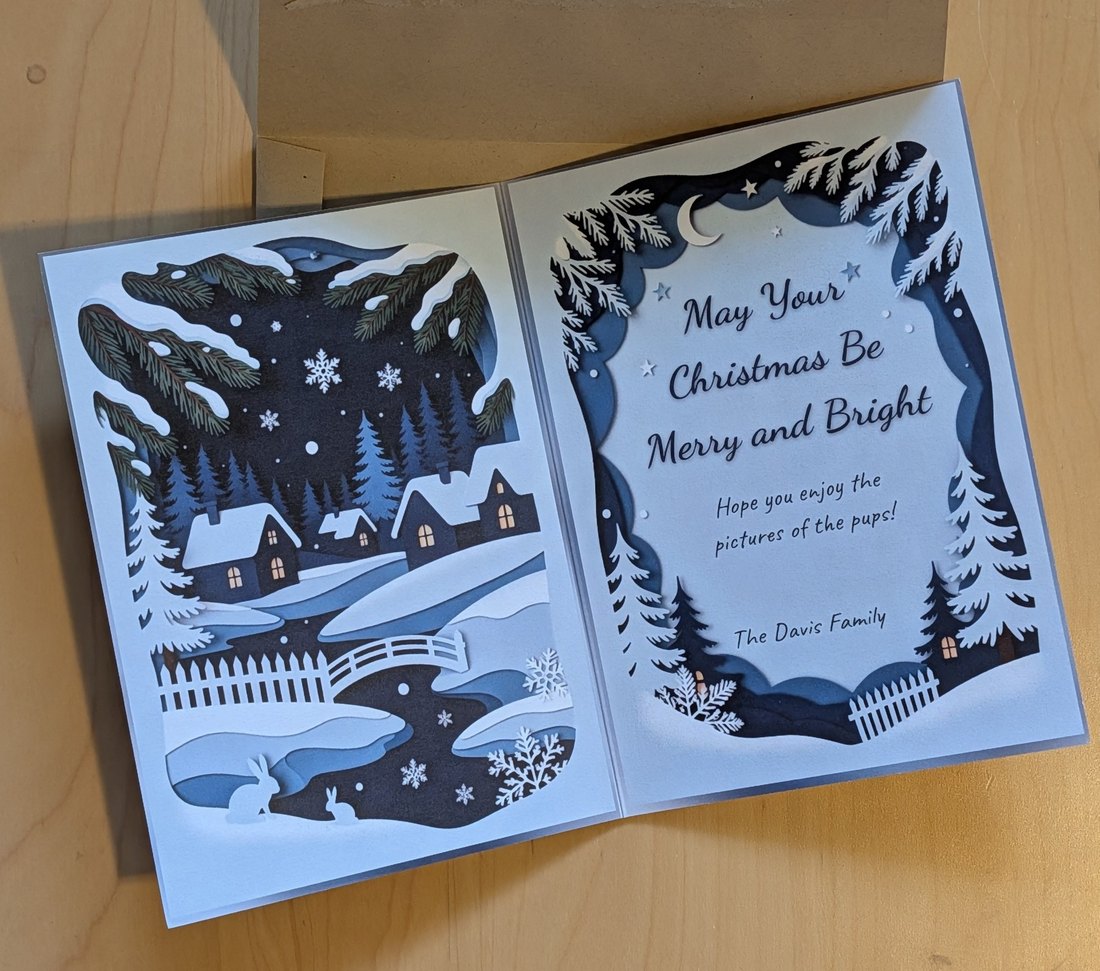 The card open on a table, a papercut winter scene on the left and a message on the right reading May Your Christmas Be Merry and Bright, Hope you enjoy the pictures of the pups, The Davis Family